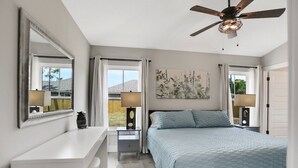 Une chambre à coucher comprenant un lit, un bureau, une chaise, un ventilateur de plafond et une fenêtre avec des rideaux.