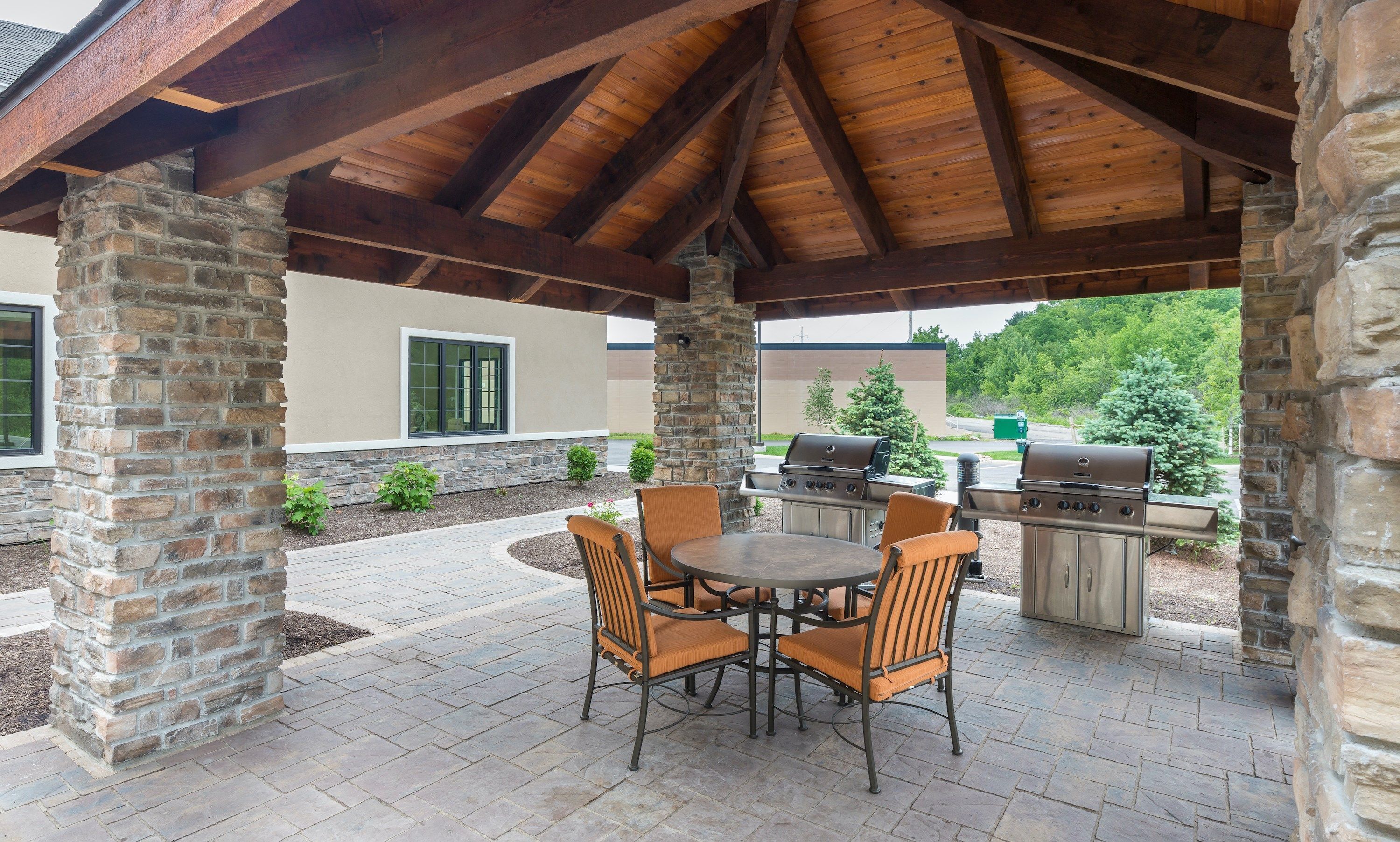An outdoor patio with stone pillars, a covered area with a wooden ceiling, a round table with chairs, and two barbecue grills.