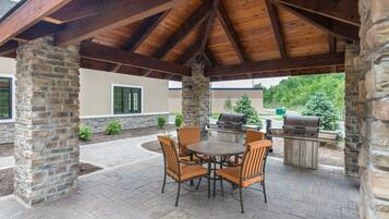 An outdoor patio with stone pillars, a covered area with a wooden ceiling, a round table with chairs, and two barbecue grills.