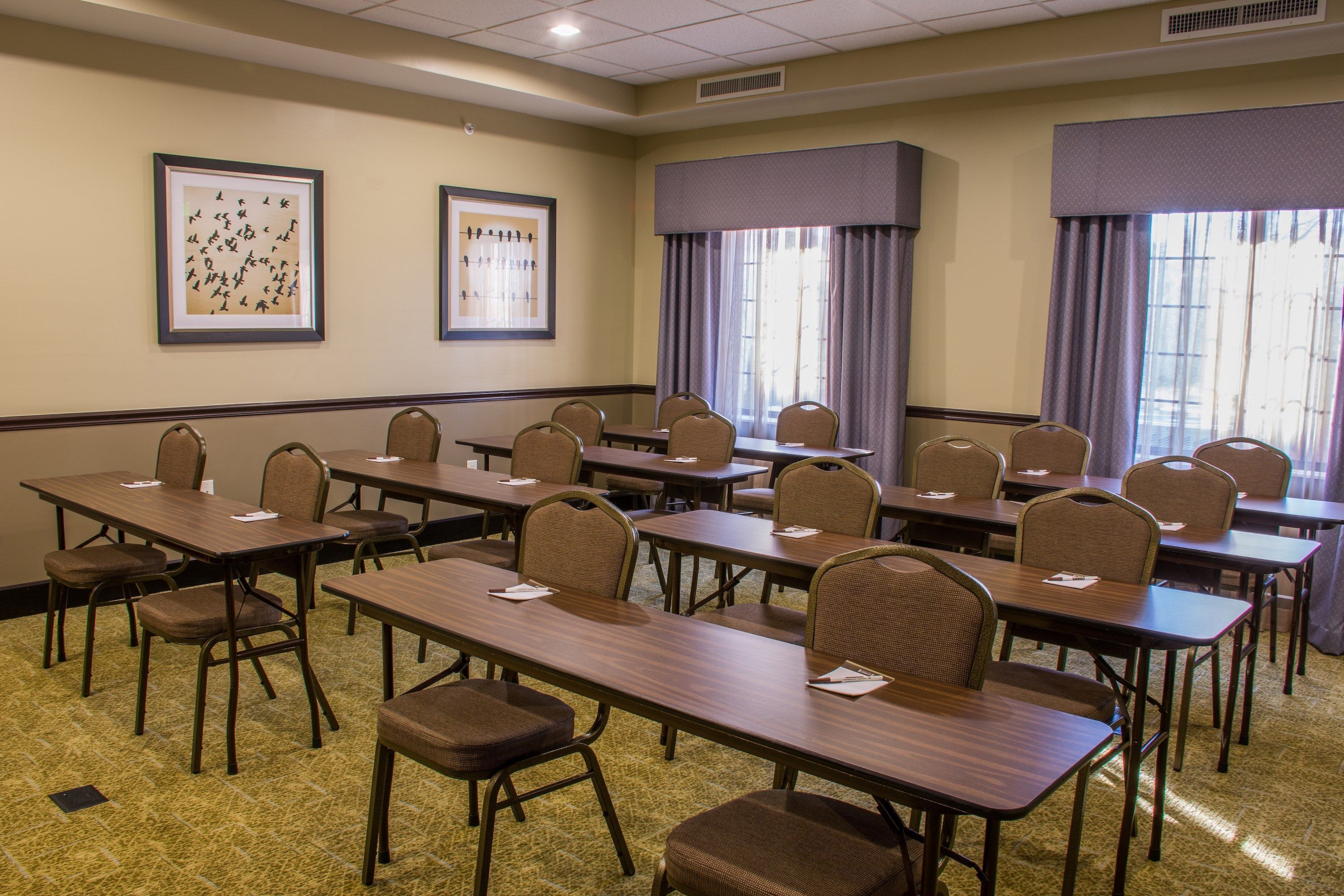 A conference room with multiple tables and chairs, framed artwork on the walls, and large windows with curtains.