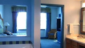 Une chambre d’hôtel avec un lit, une fenêtre avec des rideaux, une chaise et une salle de bain avec un lavabo et un miroir.