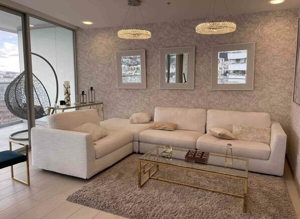 Un salon moderne avec un canapé d’angle blanc, une table basse en verre et trois tableaux encadrés accrochés au mur.