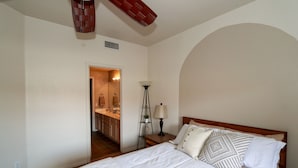 Une chambre avec un grand lit, une table de chevet avec une lampe, une tête de lit en bois, et une vue sur la cuisine et la salle de bain par une porte ouverte.