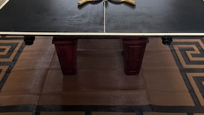 A ping pong table with two paddles and a ball in the center, a table and chairs set up outside, and a vending machine to the left.