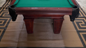 A pool table with a green felt surface, surrounded by a patterned rug, and a view of a wooded area through a glass door.
