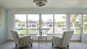 Una stanza con due poltrone decorate, un tavolino con un vaso e candele, e ampie finestre che danno su una piscina e gli edifici del resort.