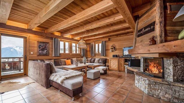 A cozy log cabin interior with a stone fireplace, wooden beams, and a large window overlooking a snowy mountain.