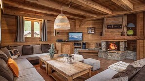 A cozy log cabin interior with a stone fireplace, a wooden ceiling, and a large window overlooking a snowy landscape.