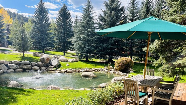 Eine Holzterrasse mit Tisch und Stühlen, einem grünen Sonnenschirm und einem Teich mit Steinen.
