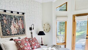 Ein Schlafzimmer mit einem großen Bett, einem Nachttisch, einer Lampe, einer Wanduhr und einem Fenster mit Blick auf Grünflächen.