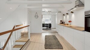 Una cocina moderna con armarios blancos, una escalera de madera y un área de estar visible al fondo.