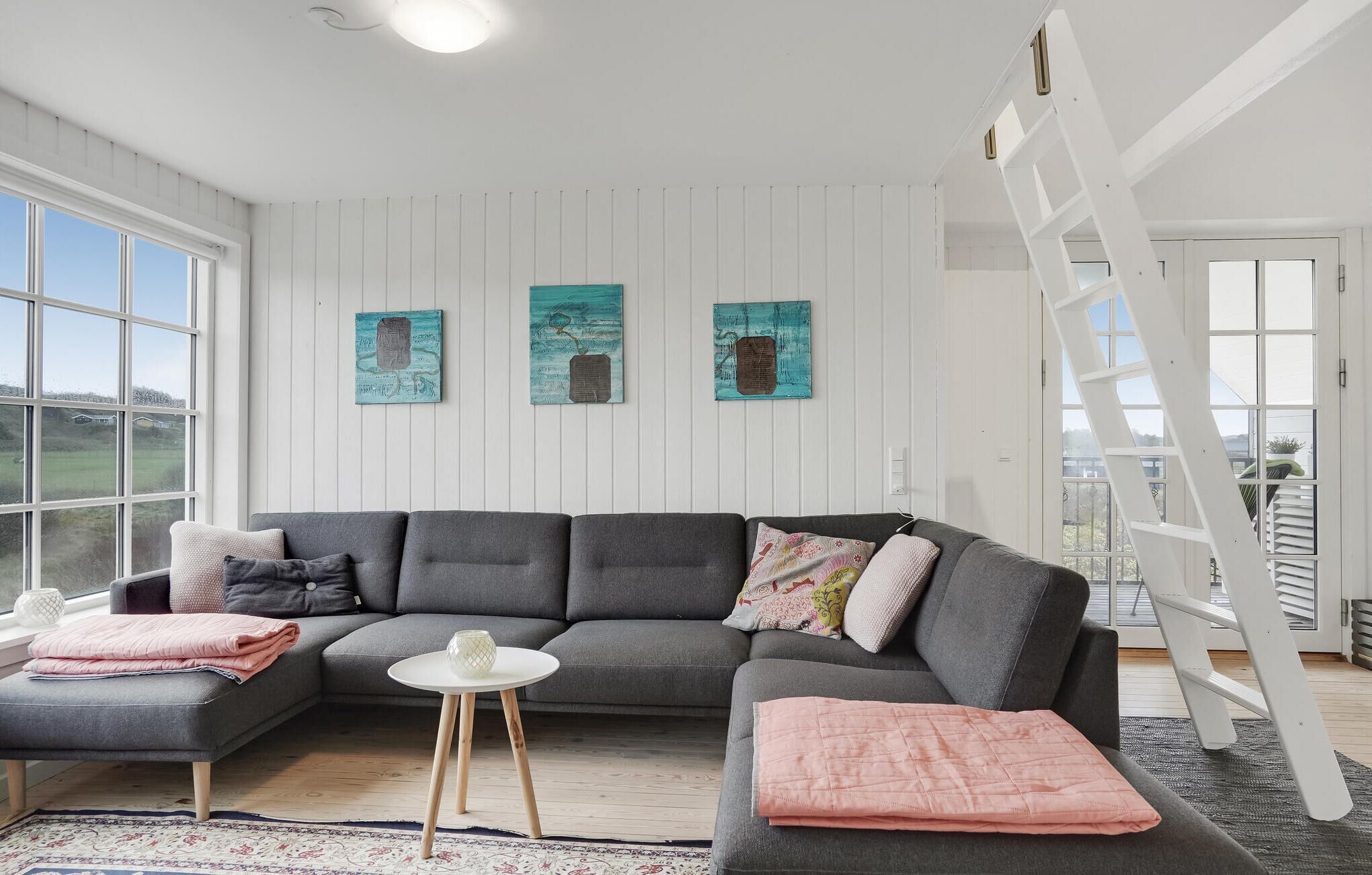 Ein modernes Wohnzimmer mit einem grauen Ecksofa, einem kleinen runden Tisch und einer Leiter. Drei Bilder sind an der Wand angebracht und ein großes Fenster bietet Blick ins Freie.