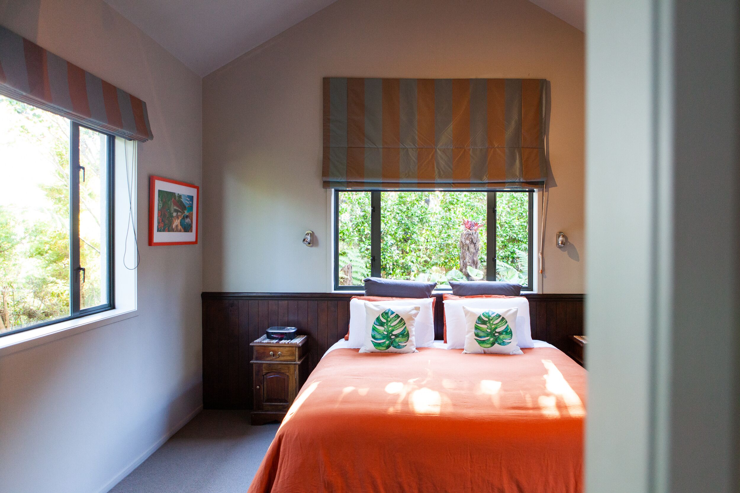 Une chambre avec une grande fenêtre, un lit recouvert d’une couverture orange et une petite table de chevet en bois.