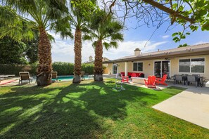 Un patio trasero con piscina, palmeras, mobiliario exterior y un porche cubierto.
