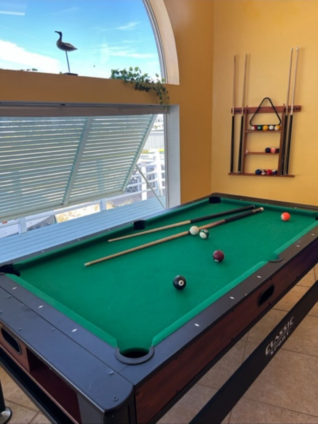 Une table de billard avec des billes, un porte-queue et une sculpture d’oiseau posée sur le rebord de la fenêtre.