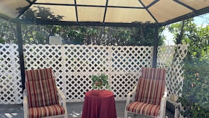 A covered patio area with two striped chairs and a table, surrounded by a white lattice fence and greenery.