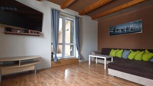 A living room with a wooden floor, a sofa with green and blue cushions, a white coffee table, a flat-screen TV mounted on the wall, and a window with curtains.