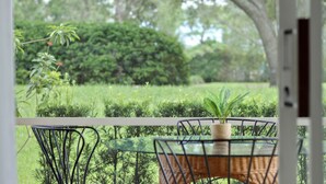A glass table with a wicker chair, a potted plant, and a view of a well-maintained garden with hedges and trees. - La Dolce Vita Studio (Port St. Lucie)