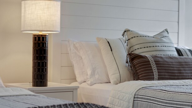 A neatly made bed with a striped blanket, a bedside lamp, and multiple pillows. - La Dolce Vita Studio (Port St. Lucie)