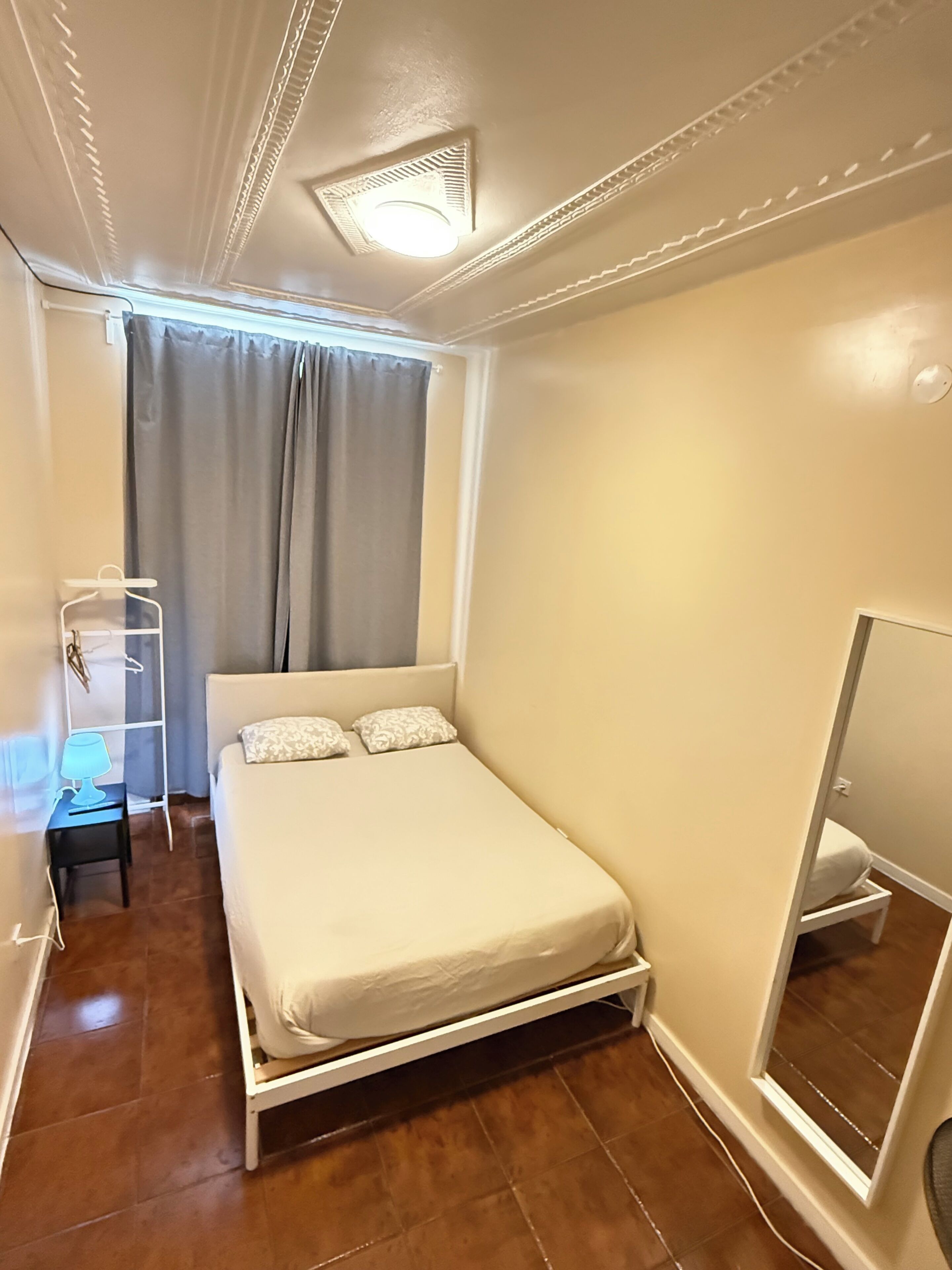 Een slaapkamer met een bed, nachtkastje met lamp, een kledingkast en een raam met gordijnen.