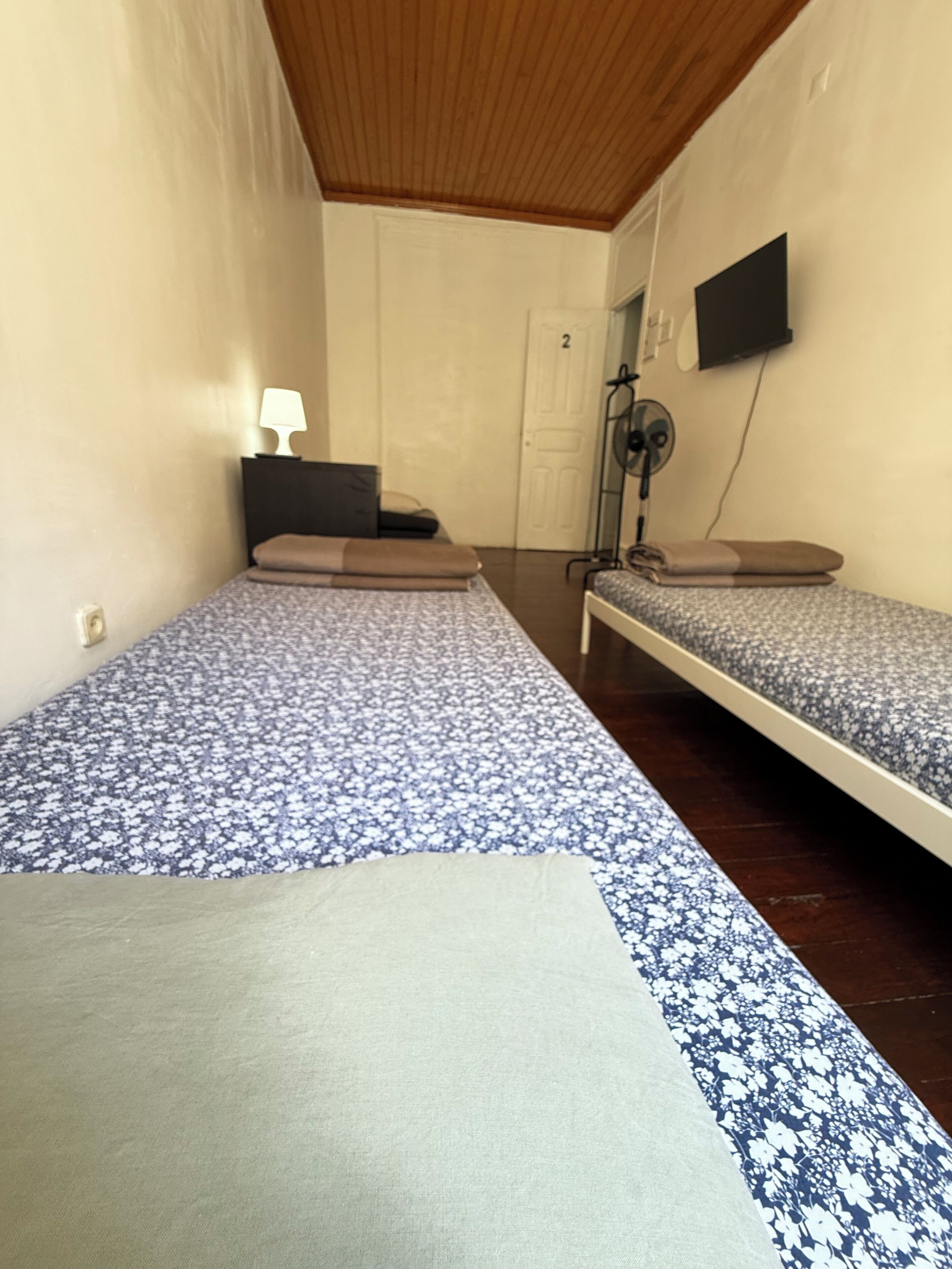 A room with two single beds, a bedside table with a lamp, a wall-mounted TV, and a door with a number.