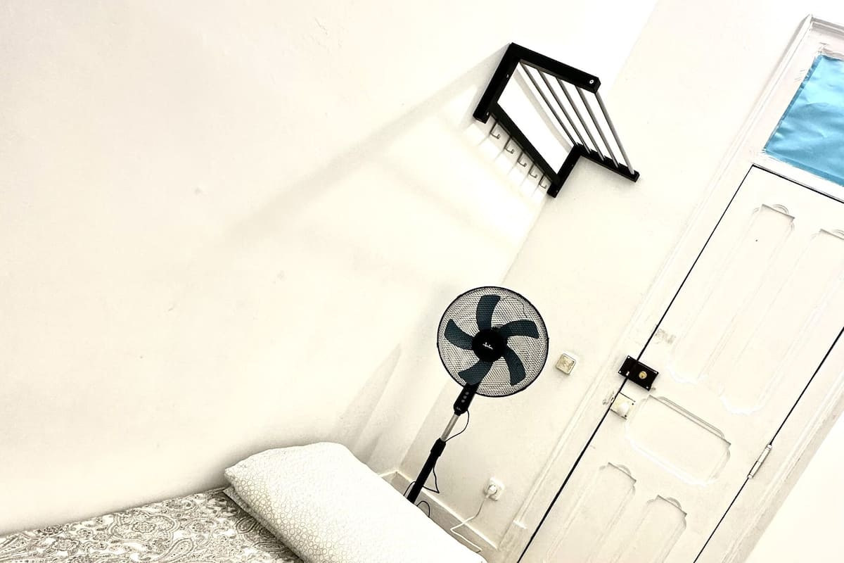 A small room with a bed, a fan, a window, and a door.