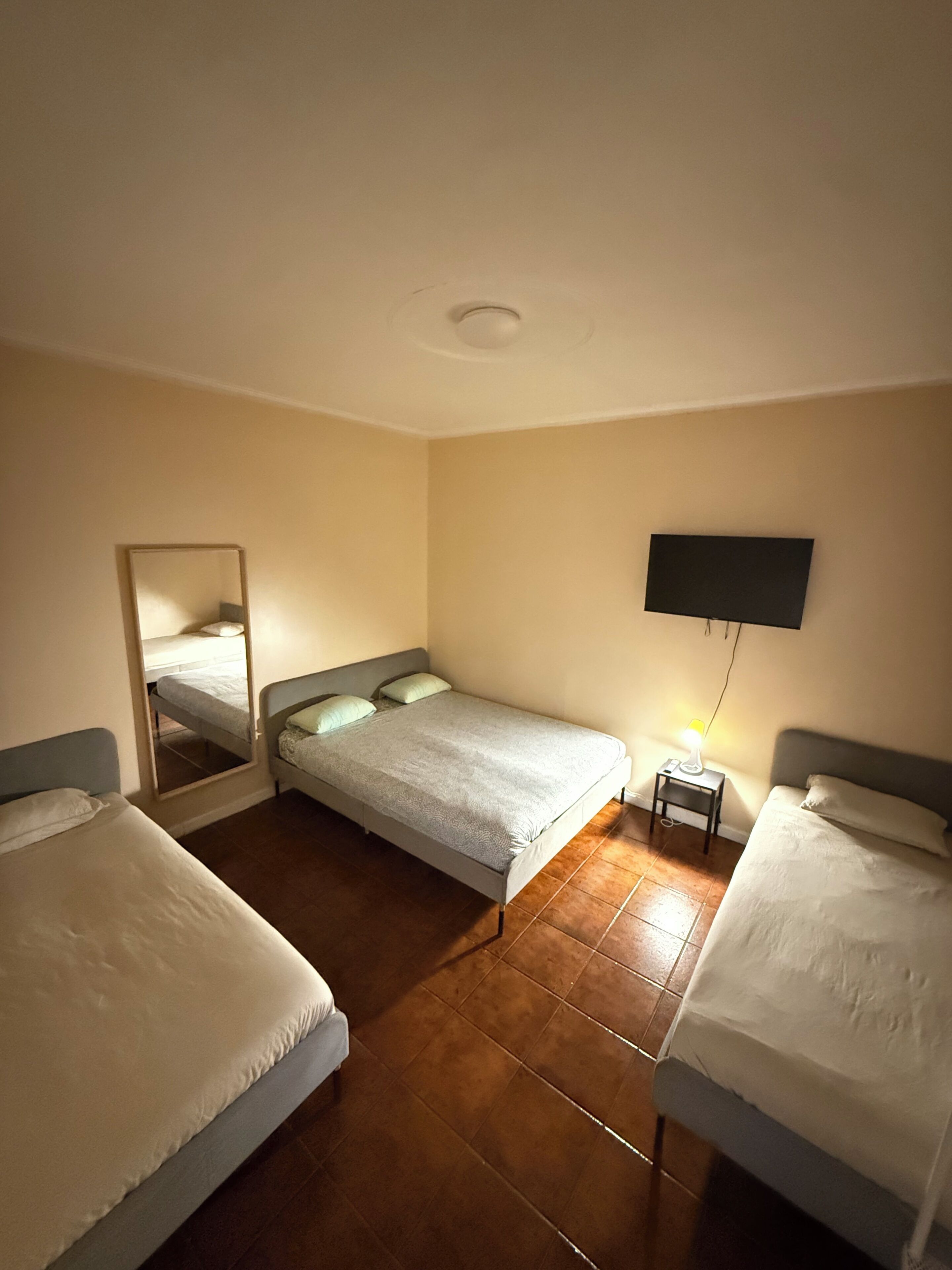 Une chambre avec deux lits simples, un téléviseur à écran plat fixé au mur, une petite table de chevet avec une lampe et un miroir sur le mur de gauche.