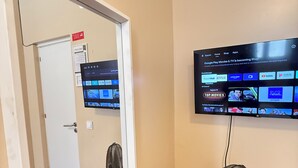 Habitación con un televisor montado que muestra la interfaz de un servicio de transmisión, una puerta que da a otra habitación y un ventilador de pie.