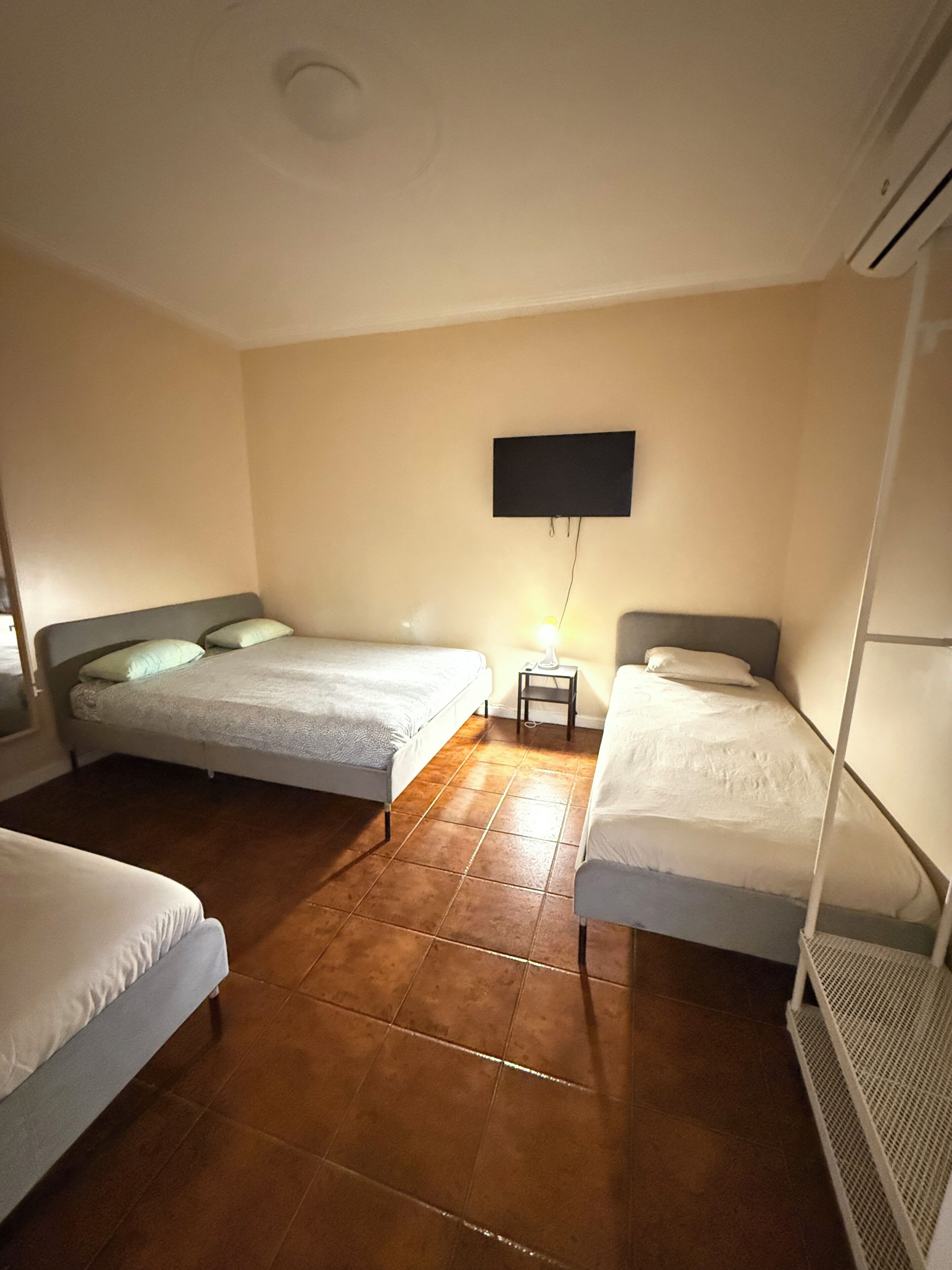 A hotel room with two beds, a flat-screen TV mounted on the wall, a small bedside table with a lamp, and a ceiling vent.