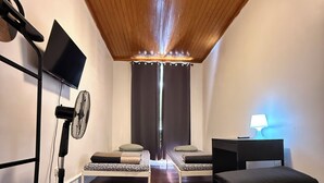 Una habitación moderna con techo de madera, dos camas, un escritorio, un ventilador de pie y un televisor.