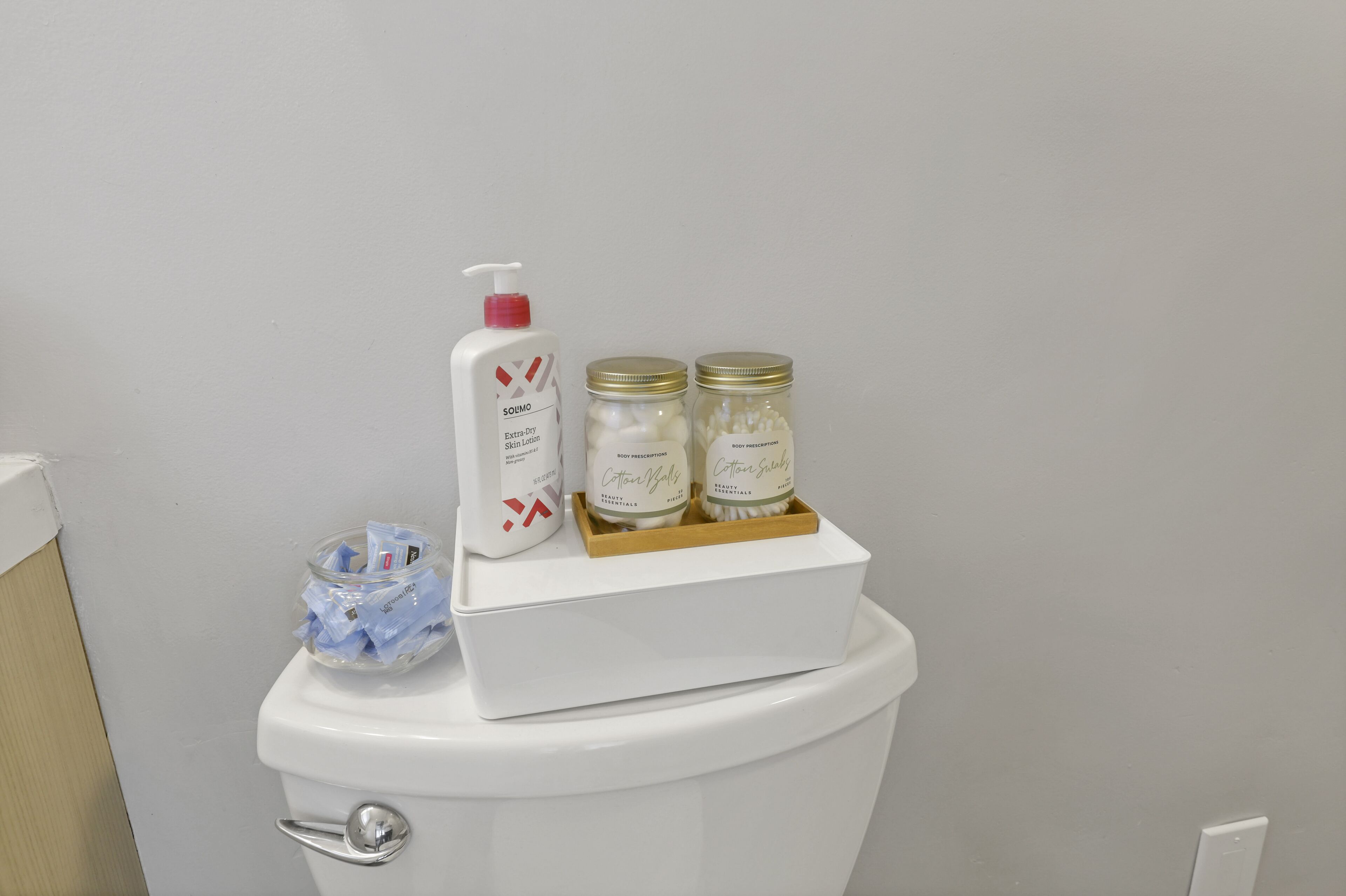 A toilet with a white lid, a bottle of hand soap, and two jars of cotton balls on top.