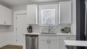 A kitchen with white cabinetry, a dishwasher, a coffee maker, and a window with blinds. - Gatsby vibes house in Old Oaks, close to everything (Columbus)