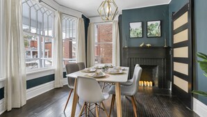 A dining area with a white table set for six, grey chairs, a black fireplace, and a view of the outside through large windows. - Gatsby vibes house in Old Oaks, close to everything (Columbus)