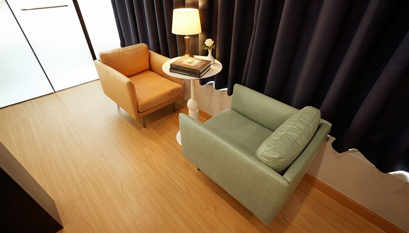A hotel room with a wooden floor, a green sofa, a beige armchair, a small round table with a lamp and a book, and dark curtains.