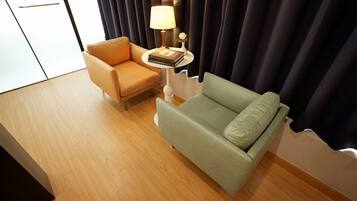 A hotel room with a wooden floor, a green sofa, a beige armchair, a small round table with a lamp and a book, and dark curtains.