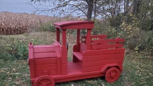 Un camioncino giocattolo rosso su un prato, con alberi e un campo di mais sullo sfondo.