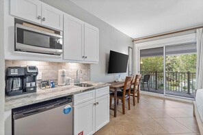 Eine moderne Küche mit weißen Schränken, einer eingebauten Mikrowelle und einer Kaffeemaschine. Ein Essbereich mit Tisch und Stühlen lädt zum Verweilen ein, und ein großes Fenster bietet Blick ins Grüne.
