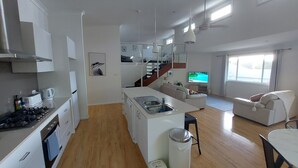 A modern kitchen with a central island, a dining area with a table and chairs, a living room with a sofa, and a TV. - Townhouse close to shelly beach Port Macquarie (Port Macquarie)