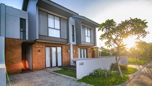 A modern two-story building with a brick facade, large windows, and a well-maintained garden area. - De Hsahe Villa (Tabanan)
