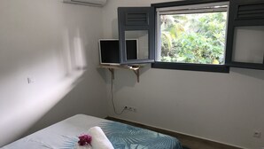 Ein Zimmer mit einem Bett, einem Fernseher auf einem Schrank, einem Fenster mit Blick auf Grünflächen und einer Klimaanlage.