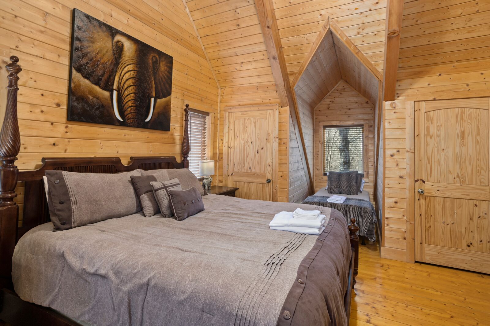 A wooden cabin bedroom with a large bed, a painting of an elephant, and a window with blinds.