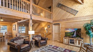 A spacious log cabin interior with a high ceiling, a balcony, leather sofas, a fireplace, and a large TV screen. - 5 Sleeping Area Log Cabin in a Great Location (Blowing Rock)