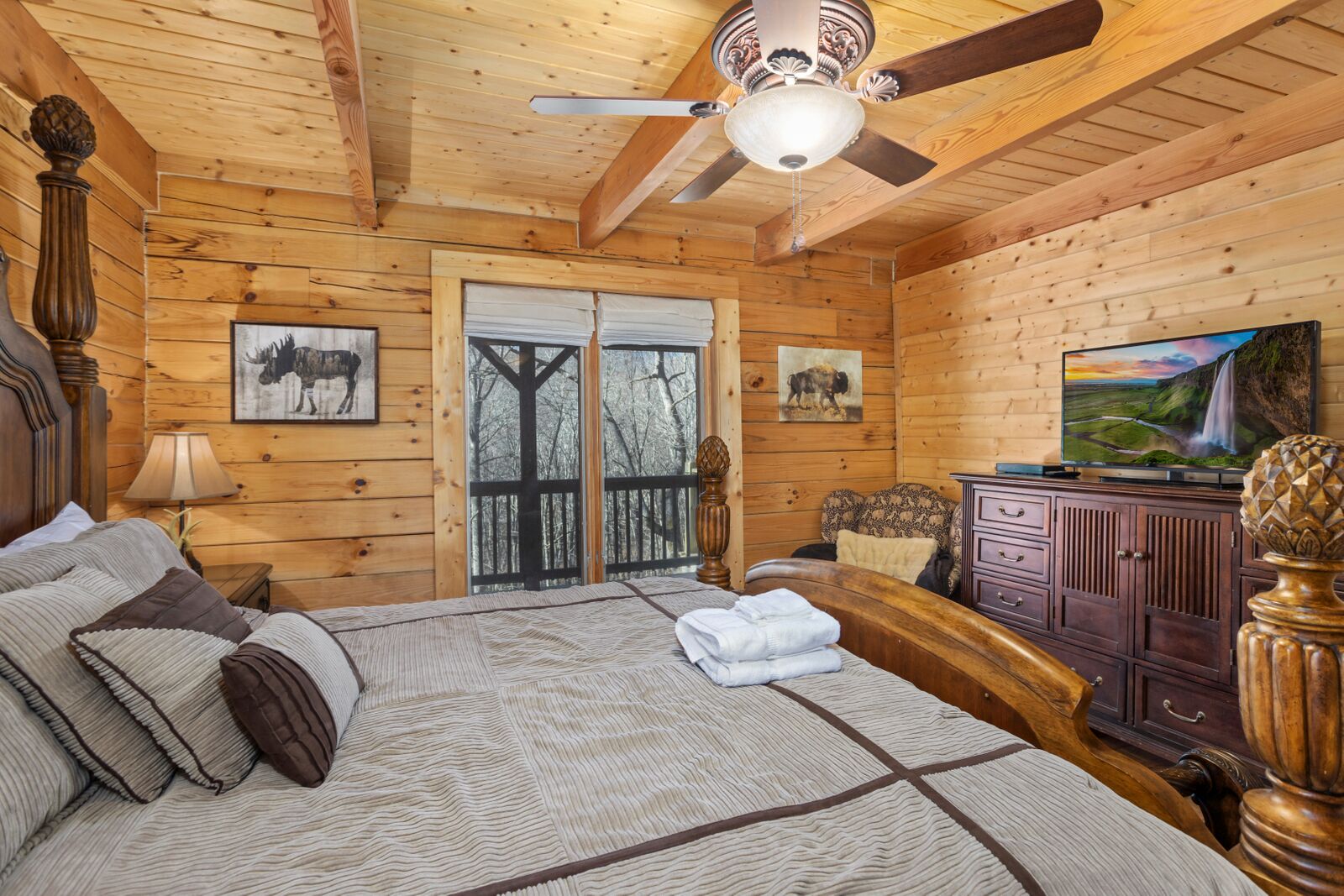 Ein Schlafzimmer in einer Blockhütte mit einem Bett, einem Schminktisch, einem Fernseher und einem Fenster mit Blick auf Bäume.