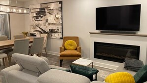 Ein modernes Wohnzimmer mit einer Couch, Sesseln, einem Couchtisch, einem Kamin, einem Flachbildfernseher und einem Essbereich mit Stühlen und einem Tisch.