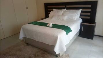 A neatly made bed with white linens and a green throw, a dark wooden headboard, a small bedside table, and a beige wardrobe.