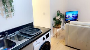 Ein kompakter Wohnbereich mit Küchenzeile, einschließlich Waschmaschine, Spüle und einer schwarzen Ablage. Es gibt ein weißes Sofa und einen Fernseher im Wohnraum.