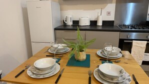 Ein Esstisch ist gedeckt mit Tellern, Schüsseln, Besteck und einem Topf mit einem Farn. Auf der Arbeitsplatte stehen eine Kaffeemaschine und ein Toaster.
