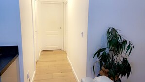 Ein schmaler Flur mit Holzboden, der zu einer weißen Tür führt. Rechts ist ein Blumentopf zu sehen.