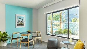 A modern room with a teal wall, a round table with chairs, a potted plant, a window with a view of palm trees and mountains, and a small side table with a book and vase. - Twin Palms Resort - A Gay Men's Resort (Palm Springs)