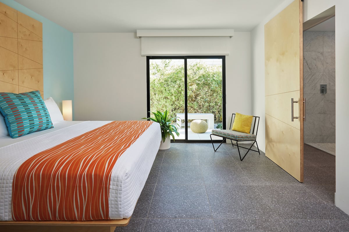 Een moderne hotelkamer met een groot bed, een kamerplant, een stoel en uitzicht op het groen door een schuifdeur.